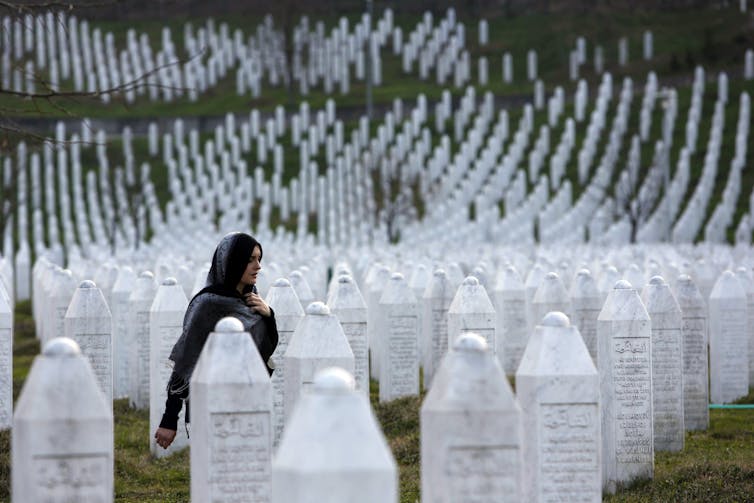 A woman with a scarf on her head walks in a cemetery among a sea of white gravestones.