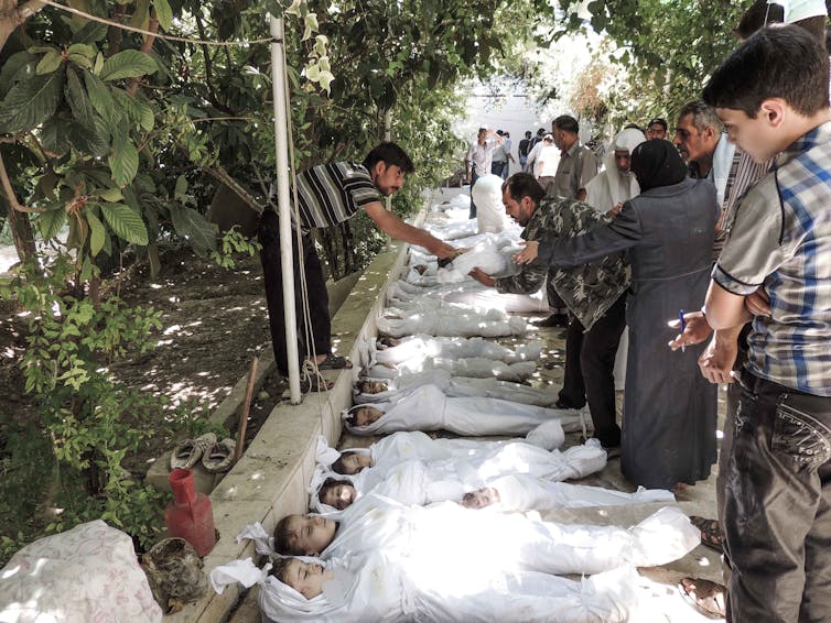 A row of dead children, covered in white cloth, is shown, as adults look over them