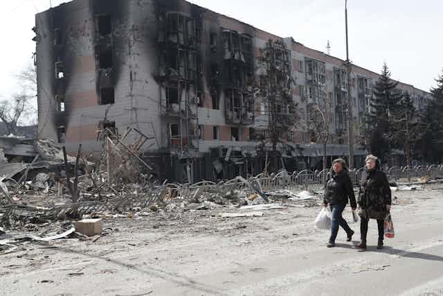 Two women walk, wearing dark clothing, in front of debris and a charred building.