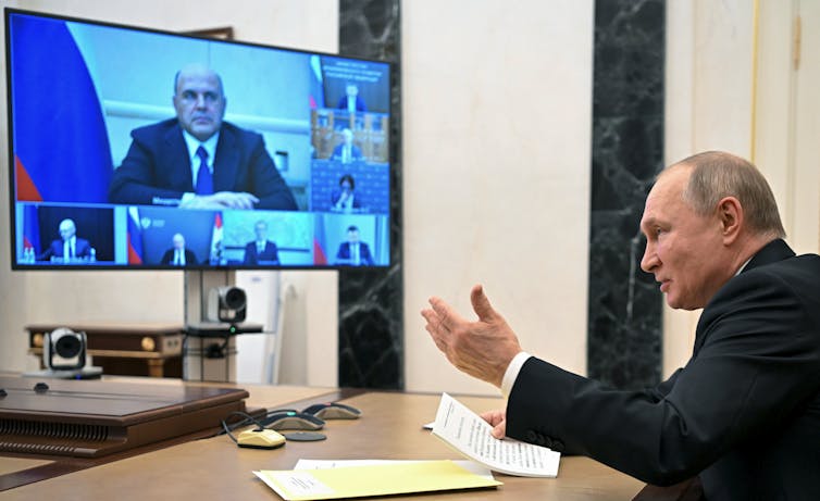 A white balding man gestures with his left hand over desk as he looks at a monitor that shows one white man in a big window and others around him