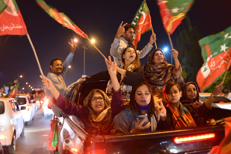 Women and men holding flags in the back of a truck cheer and gesture.