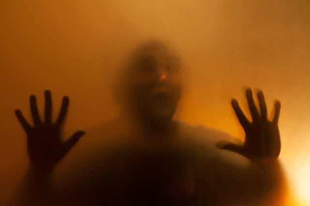 A shadowy image of a man behind a translucent facade with hands pressed up against glass. 