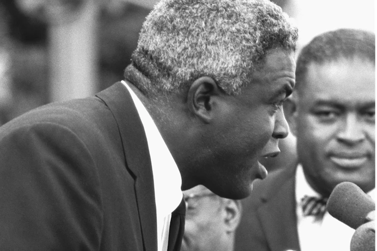 Black man with white hair speaks into a microphone.