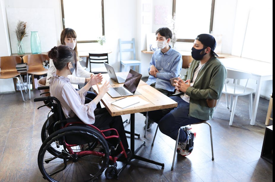 Four people wearing facial masks sit at a table together. One of the women is in a wheelchair and she is gesturing with her hands as she speaks.