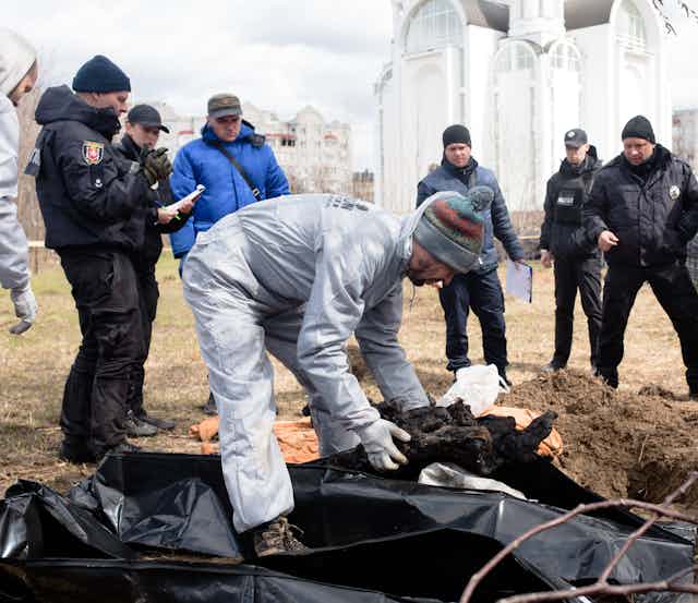 A man wearing gloves looks at several dead bodies covered in dark plastic.