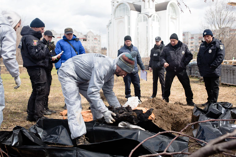 A man wearing gloves looks at several dead bodies covered in dark plastic.