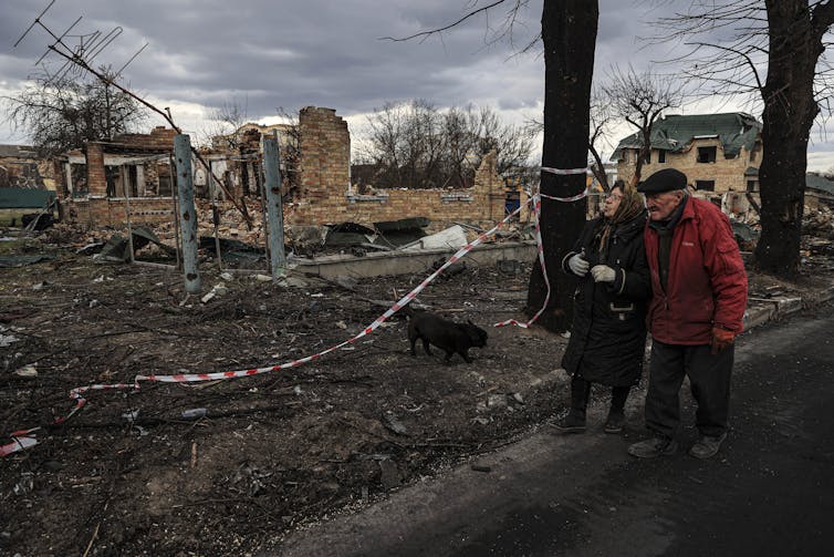 An elderly man and woman are seen walking through rubble near a destroyed house.