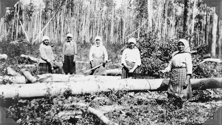 Black and white photo from about the turn of the last century (1900) of two women in traditional Ukrainian dress cutting logs.