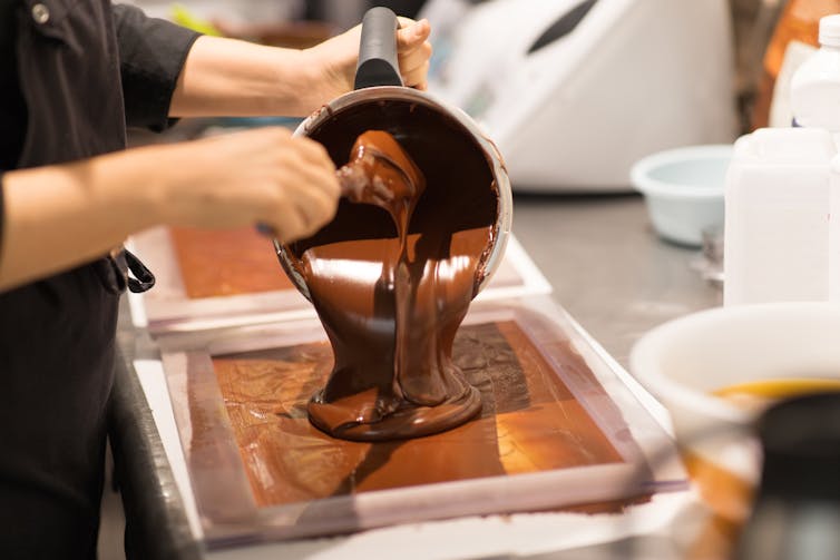 A photograph of a person pouring molten chocolate from a pot into a tray.