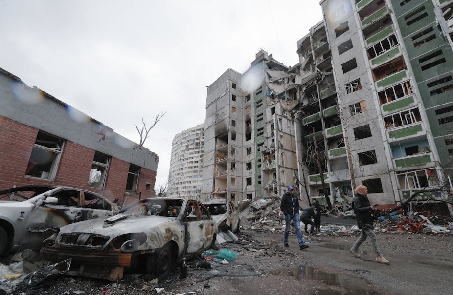 A women walks ahead of a man as they pass apartment buildings and cars destroyed by Russian bombs.