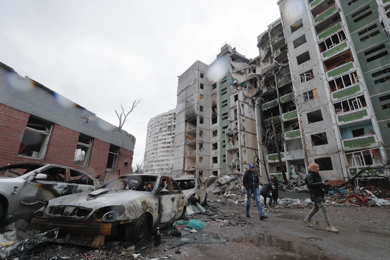 A women walks ahead of a man as they pass apartment buildings and cars destroyed by Russian bombs.
