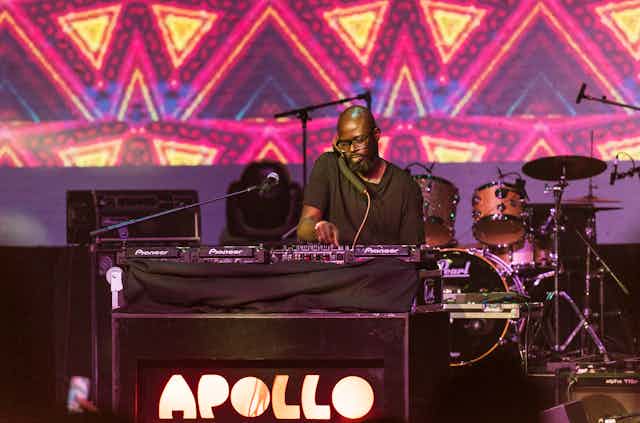 Bright African-patterened lights in gold and pink shine behind a man deejaying in a plain black T-shirt, wearing glasses and a beard with a shaved head.