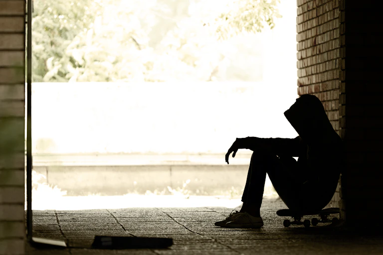 Young boy sits on skateboard, hunched over and in the dark.