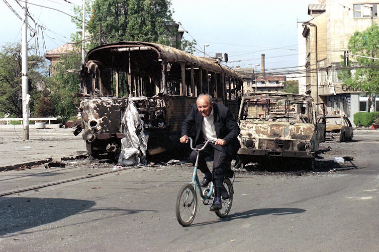 An elderly man rides his bike past the burnt out wreckage of a bus and cars.