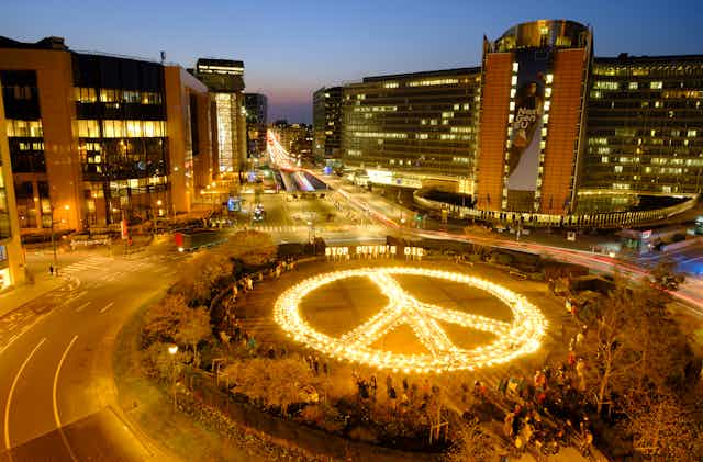 A giant peace sign is lit up with the sign Stop Putin's Oil behind it in a city plaza