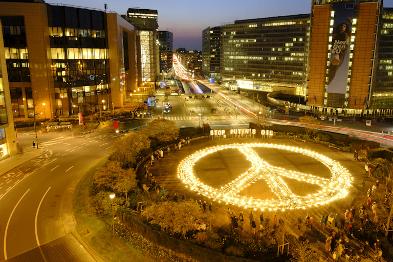 A giant peace sign is lit up with the sign Stop Putin's Oil behind it in a city plaza