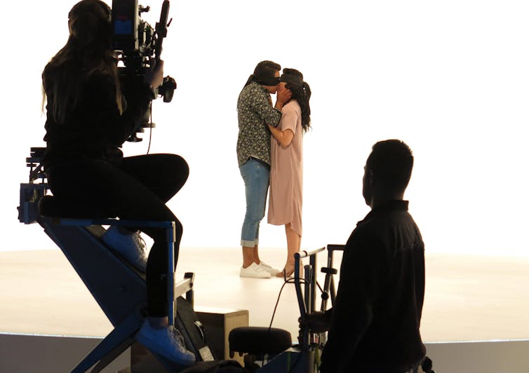 Two blindfolded people kissing while cameras film them