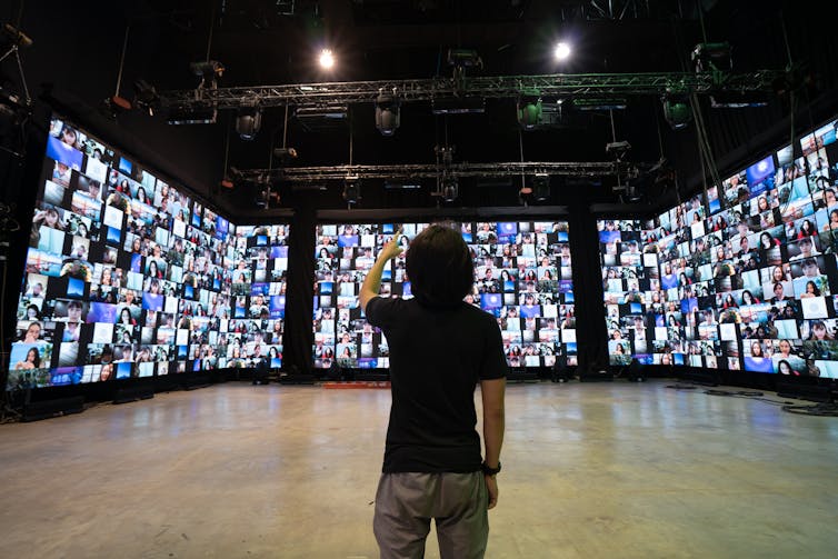 A man in grey jeans and a black t-shirt stands in a large room in front of three tall screens filled with hundreds of zoom windows.