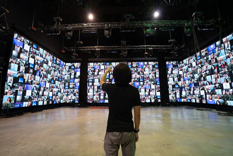 A man in grey jeans and a black t-shirt stands in a large room in front of three tall screens filled with hundreds of zoom windows.