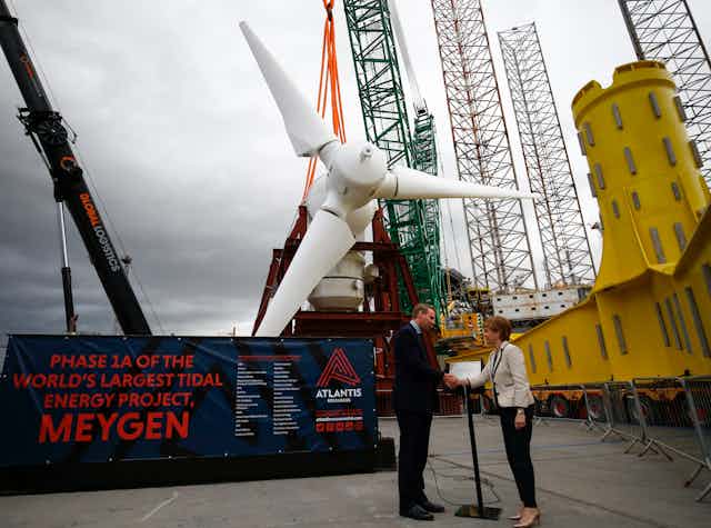 Two people shake hands in front of a large white turbine