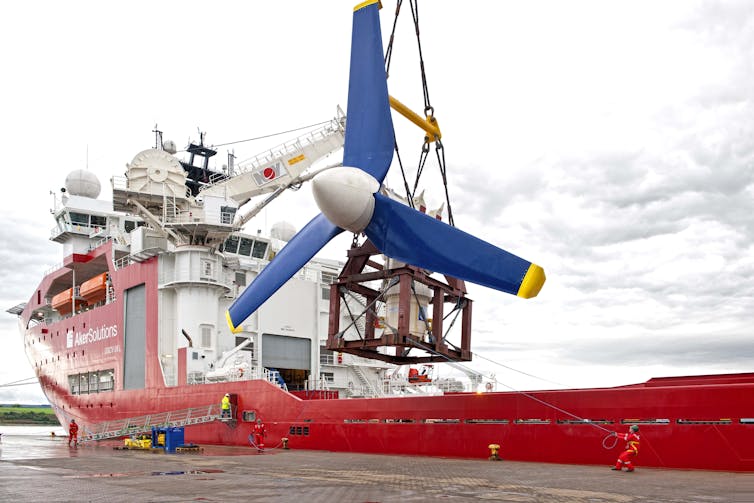 A large turbine with blue blades is suspended from a ship