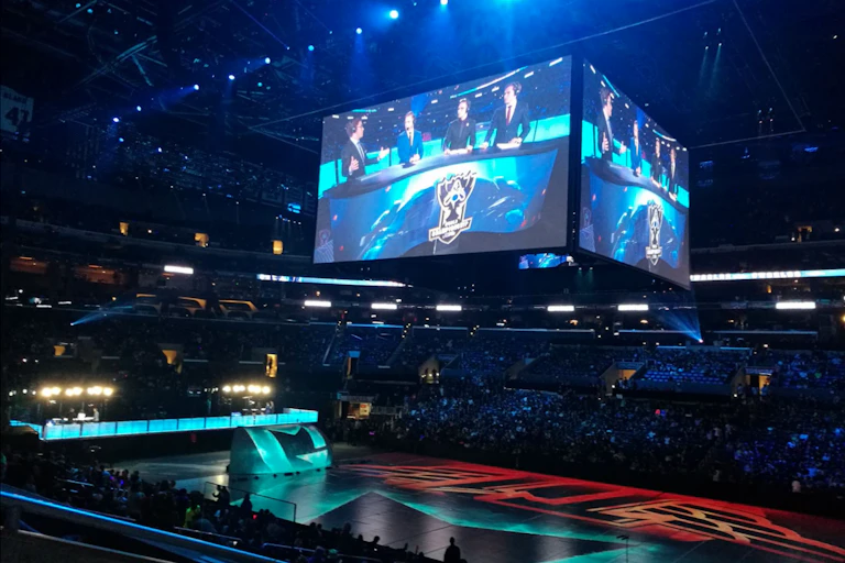 A brightly lit stadium with a huge screen showing a panel of four commentators.