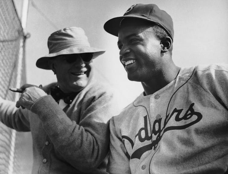 A white man in a large hat is seen talking with a Black baseball player wearing a Brooklyn Dodgers uniform.