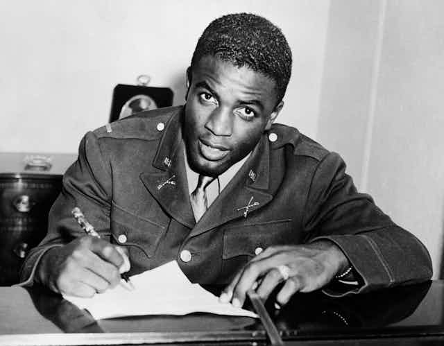 A black man wearing a military uniform is signing a sheet of paper as he stares into the camera.