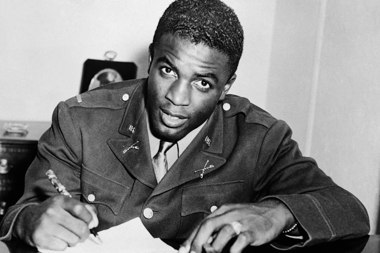 A black man wearing a military uniform is signing a sheet of paper as he stares into the camera.