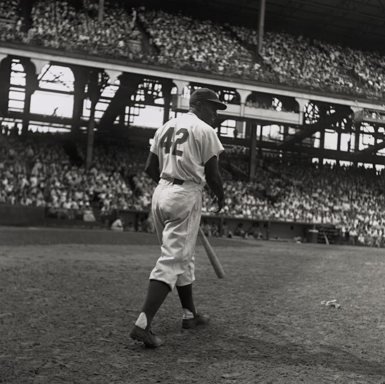 A Black baseball player wearing the number 42 steps up to bat in a stadium packed with thousands of fans.
