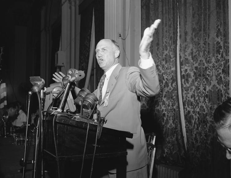 A white man dressed in a business suit is standing behind a lectern and gesturing with one hand.