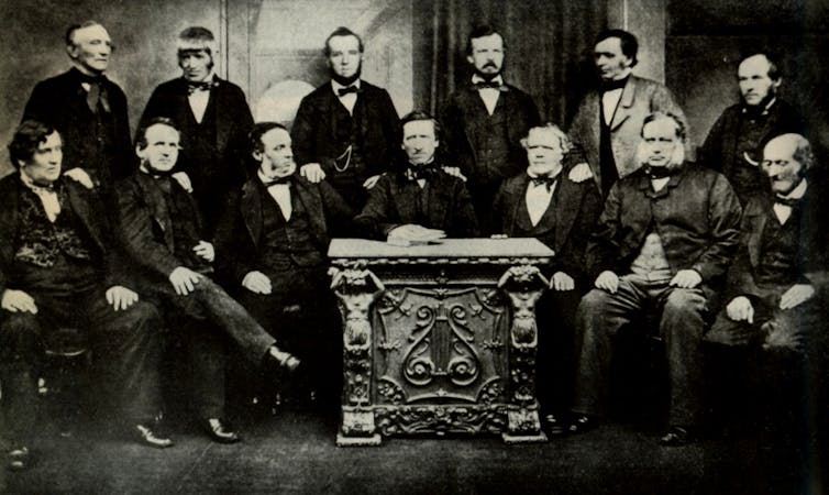 A black and white photo show a group of men sitting around a table.