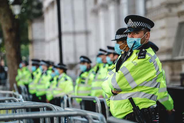 A row of policemen and women in black hats and high-vis vests and masks stand on a pavement behind metal barriers.