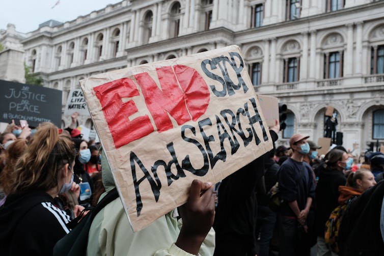 A crowd of protestors in a street setting hold up signs against stop and search.