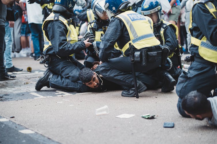 Two men are held flat on the ground by a group of police offiers in riot gear.