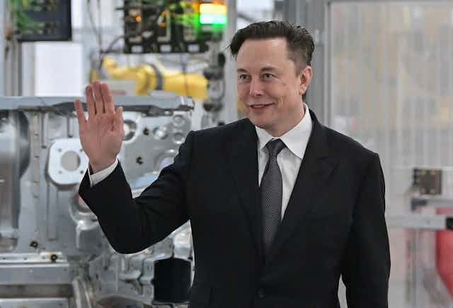 A white man in a black suit holds his right up as he smiles into the distance with machinery in the background