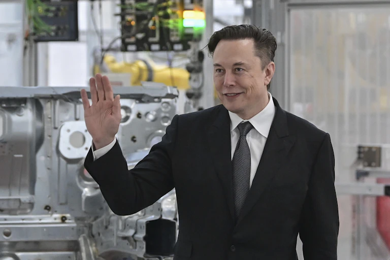 A white man in a black suit holds his right up as he smiles into the distance with machinery in the background