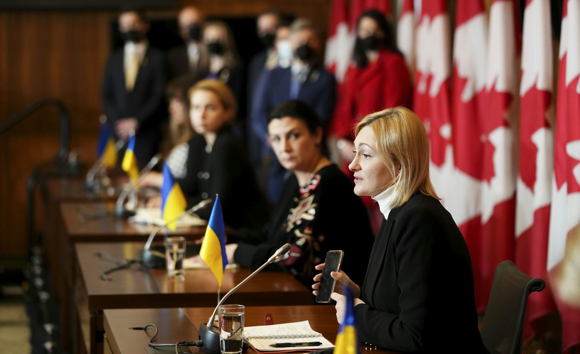 A blonde woman speaks at the end of a table with other women sitting beside her. Small Ukrainian flags are in front of them and a row of large Canadian flags are behind them.