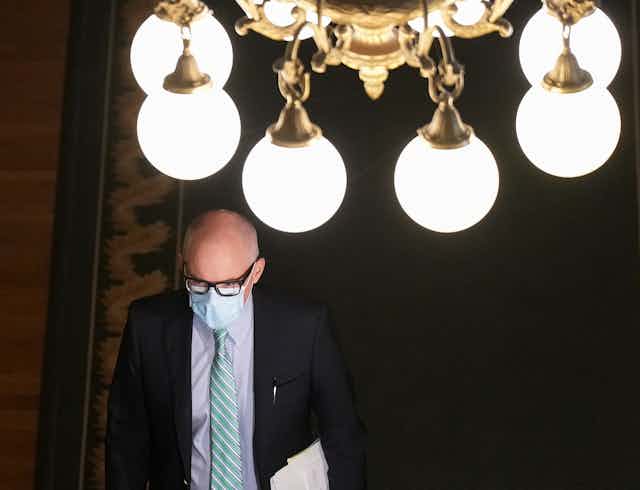 A man in a suit and tie, wearing a face masks, walks under a light fixture with several large white globes