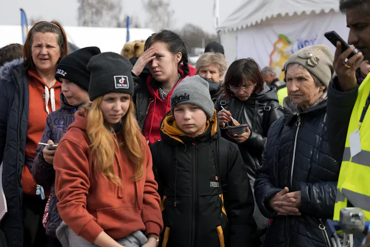 A girl with red hair, wearing a black cap and orange jacket, stands next to a boy in a black knit cap among several other people at a border crossing.