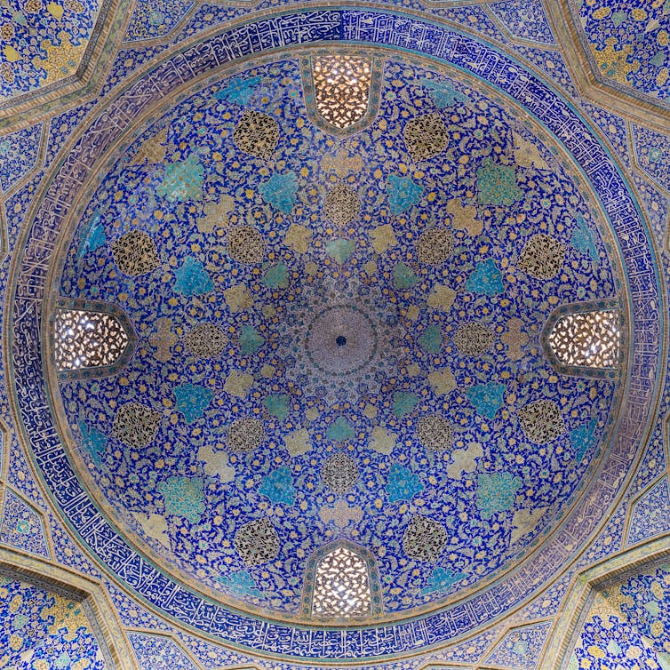 Highly highly detailed ornament, in hues of gold, tan, teal and brown, is seen organized in a circular patterns, is seen on a cobalt blue cieling.