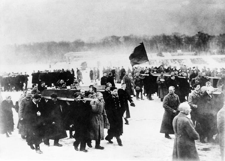 A black and white photo shows groups of men in dark coats carrying caskets on their shoulders in a snowy scene. Someone in distance holds a flag