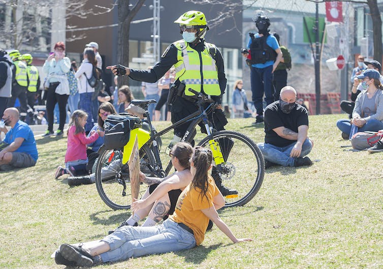 Un policier à vélo parle avec des gens assis par terre, dans un parc