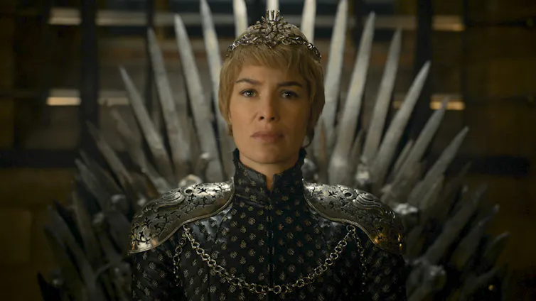 A white woman wearing a crown and dark clothing that goes up above her neck sits on a throne made of swords