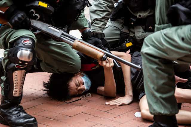 A man wearing a mask is shown pushed down on the floor, surrounded by police with large guns and wearing green uniforms