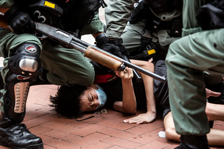 A man wearing a mask is shown pushed down on the floor, surrounded by police with large guns and wearing green uniforms