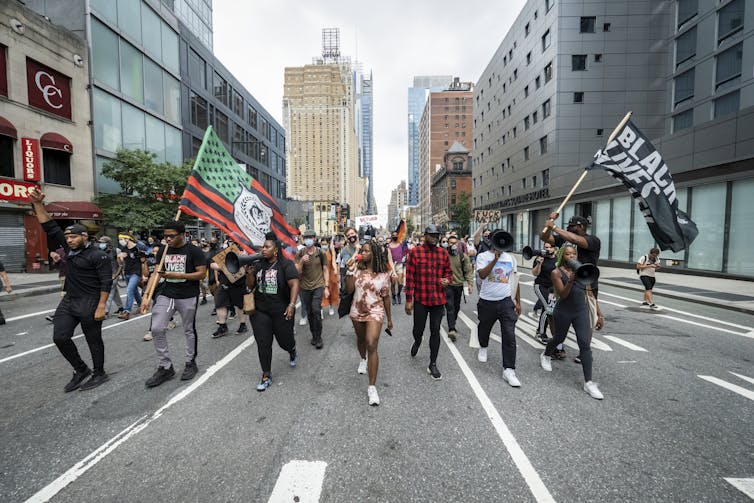 Rows of young people, some of whom are holding Black Lives Matter flags, walk together down an empty Manhattan street