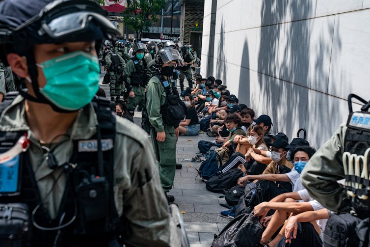 Police wearing face masks stand over a row of young people seated against a wall in Hong Kong.