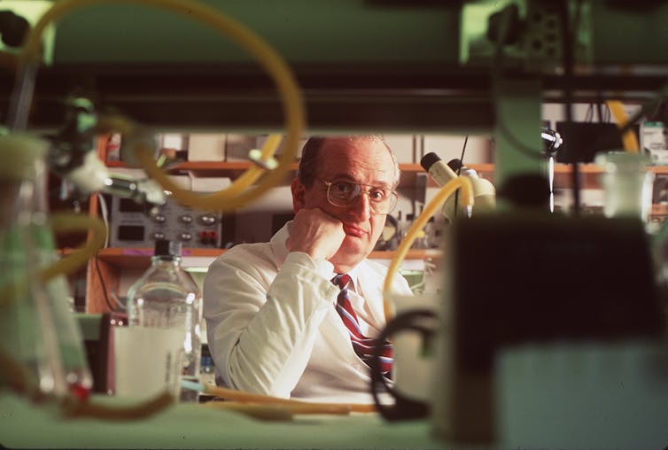 A balding man with glasses wearing a lab coat over a white shirt and tie sits at a lab bench surrounded by test tubes and lab equipment.