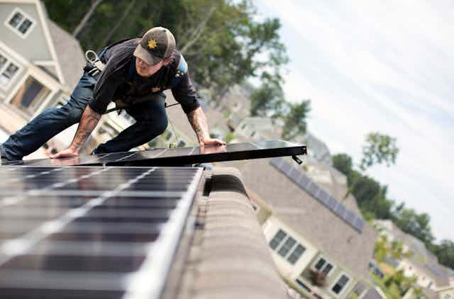 A man installs solar panels on a roof with other solar homes behind him